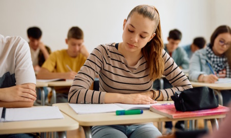 Reading literacy: Teen girl with long blonde hair in ponytail sits in classroom reading book on her desk with other students in background reading at their desks