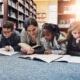 Book Ban Survey: Several young elementary students with one adult female teacher lie on library blue rug reading books in front of bookshelves filled with books