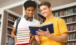 Teach literature classics: Two teen boys stand next to each other in front of full bookcases. One holds a stack of books while the other holds an open book they are both reading.