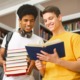 Teach literature classics: Two teen boys stand next to each other in front of full bookcases. One holds a stack of books while the other holds an open book they are both reading.