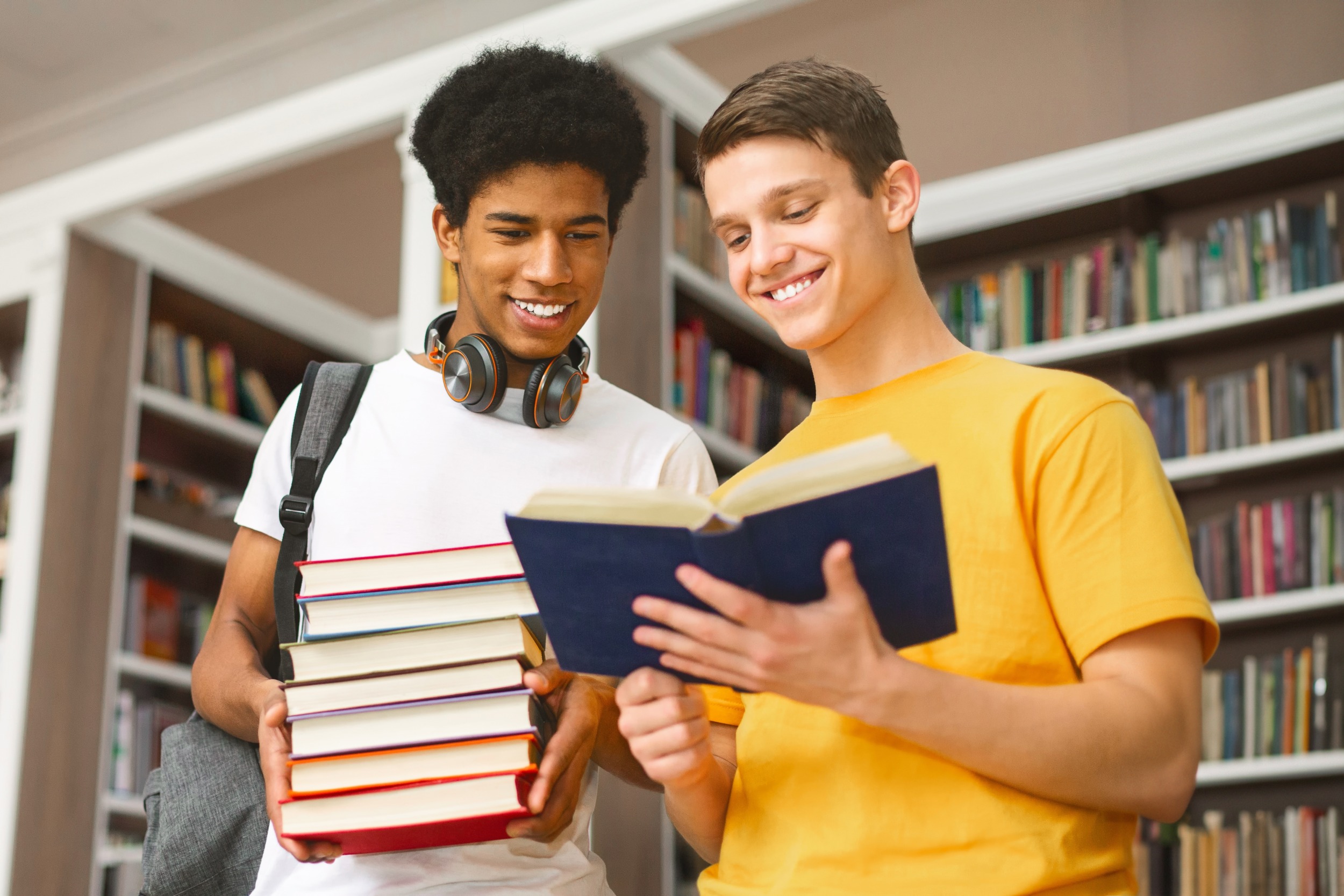 Teach literature classics: Two teen boys stand next to each other in front of full bookcases. One holds a stack of books while the other holds an open book they are both reading.