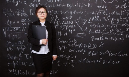 Teaching scientific theory: woan teacher with dark hair pulled back in black jacket, white shirt and black skirt, holds black folder standing in front of black board completely filled with white chalk science formulas.