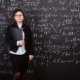 Teaching scientific theory: woan teacher with dark hair pulled back in black jacket, white shirt and black skirt, holds black folder standing in front of black board completely filled with white chalk science formulas.