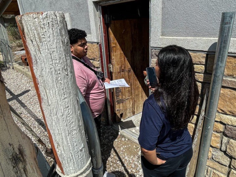 Reduce chronic absenteeism: Woman with long dark hair, navy top and black pants stands outside a house talking on a cell phone, facing a man with dark hair, pink t-shirt and blue jeans.