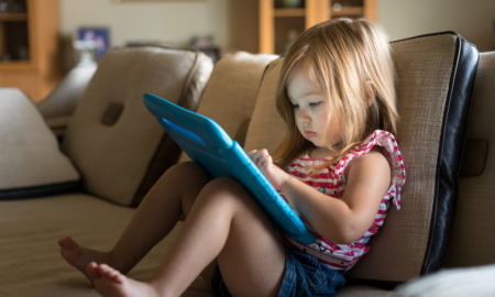 Preschooler excessive screen time study: Young girl in red top and blue shorts with long light brown hair sitting at home on tan couch and using a child's tablet touch screen computer