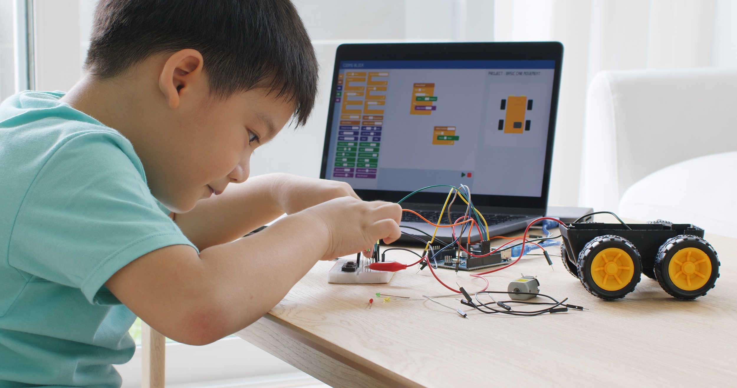 education opportunity gag: Young boy with dark hait sits at desk playing with wiring on robot next to a computer monitor