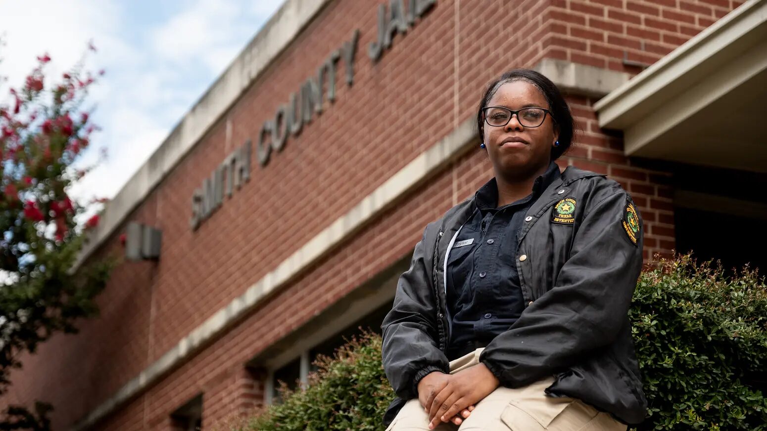 Teens as Texas Jail Guards: Teen girl in tan pants and dark uniform jacket sitting outside a red brick building on a low red brick entryway wall