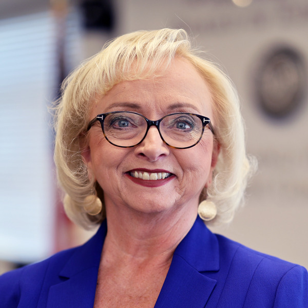 Edu R&D Budget: Head shot older woman with bobbed blonde hair, dark-framed glasses in Mediterranean blue jacket