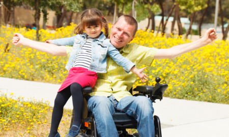 Disabled parenting: Man in wheelchair with short hair in light shirt and blue jeans holds young dark-haired girl in blue shirt, red skirt and black tights sitting on the arm of hi wheelchair