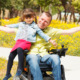 Disabled parenting: Man in wheelchair with short hair in light shirt and blue jeans holds young dark-haired girl in blue shirt, red skirt and black tights sitting on the arm of hi wheelchair