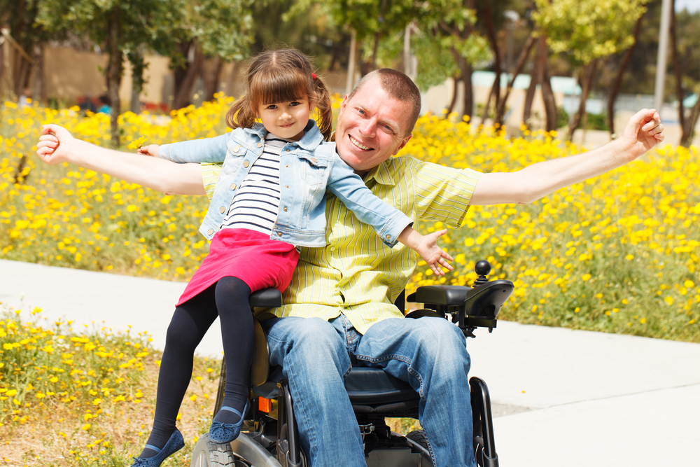 Disabled parenting: Man in wheelchair with short hair in light shirt and blue jeans holds young dark-haired girl in blue shirt, red skirt and black tights sitting on the arm of hi wheelchair