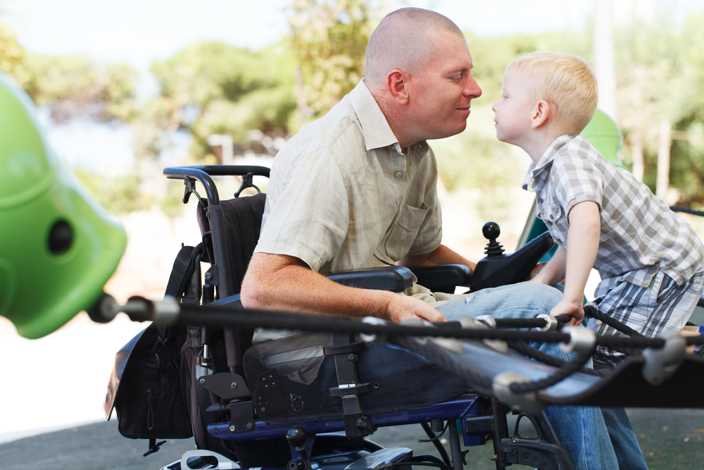 Disabled parents: Man in wheelchair in light shirt and blue jeans leans forward to wards a young boy with blonde hair in plaid shirt and shorts who is standig on the wheelchair feet rest and holding onto arms of wheelchair