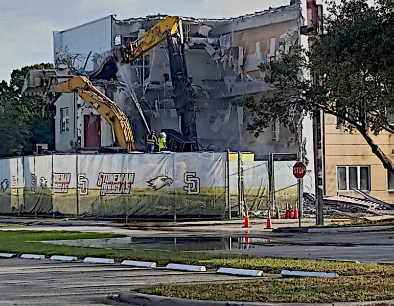 School shooter drills: Three story modern building under demolition by two large, pieces of heavy machinery, all behind a temporary wood fence painted with a grassy filed and several instances of Stoneman Douglas name and logo.