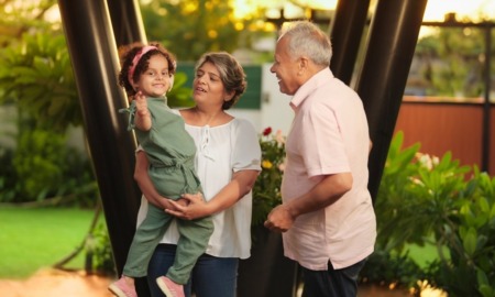 Kinship foster care: One older man and one older woman both with gray hair stand outside with grennery in front of green trees, the woman holding a dark-haired, female todder in sage green jumpsuit on her hip as girls waves at camera.