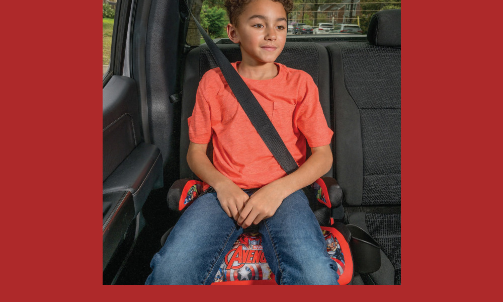 report child booster seat safety: Young elementary-age boy with short, dark, curly hair, orange t-shirt and bluejeans, sits in booster eat strapped into back seat of vehicle with black upholstery