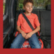 report child booster seat safety: Young elementary-age boy with short, dark, curly hair, orange t-shirt and bluejeans, sits in booster eat strapped into back seat of vehicle with black upholstery