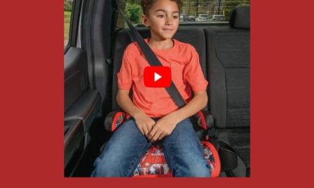 report child booster seat safety: Young elementary-age boy with short, dark, curly hair, orange t-shirt and bluejeans, sits in booster eat strapped into back seat of vehicle with black upholstery