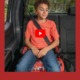 report child booster seat safety: Young elementary-age boy with short, dark, curly hair, orange t-shirt and bluejeans, sits in booster eat strapped into back seat of vehicle with black upholstery