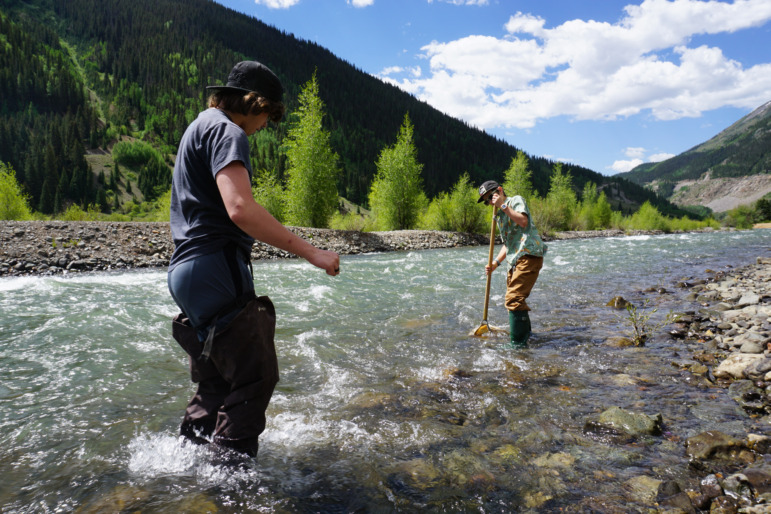 Rural school districts join forces: Two teen boys standin a shallow river wearing water gators gathering ecosystem samples from the water