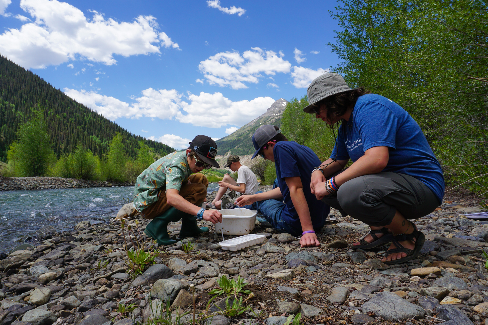 Rural school districts join forces: Three teens and a male adult sit on rocks of river shore gathering ecosystem samples