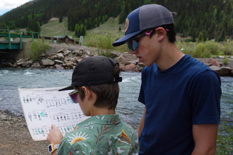 Rural school districts join forces: Two teen boys stand together next to a river marking a chart one boy holds