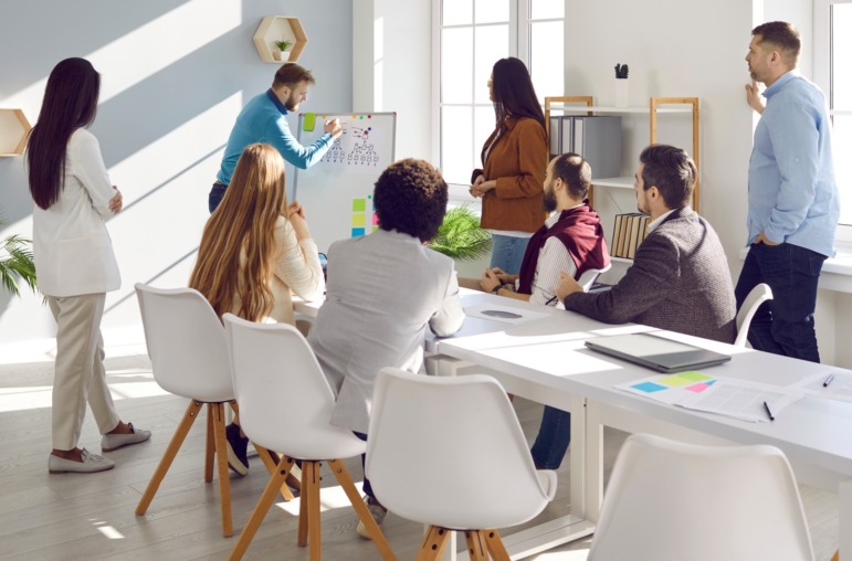 AI replace teachers: Group of adult students stand and sit at long white table intently watching adult male at drawing and explaining diagram on white board standing at front of table.