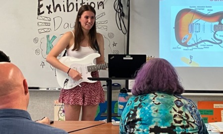 Competency-based innovative high school: Teen girl with long dark hair in white top and red print skirt stands with white guitar in front of classroom singing a song