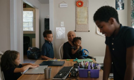 Online schools: 4 children with dark hair sit and stand around a dining table with school supplies in front of each of them while a woman in hijab leaning down next to one of them