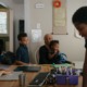 Online schools: 4 children with dark hair sit and stand around a dining table with school supplies in front of each of them while a woman in hijab leaning down next to one of them