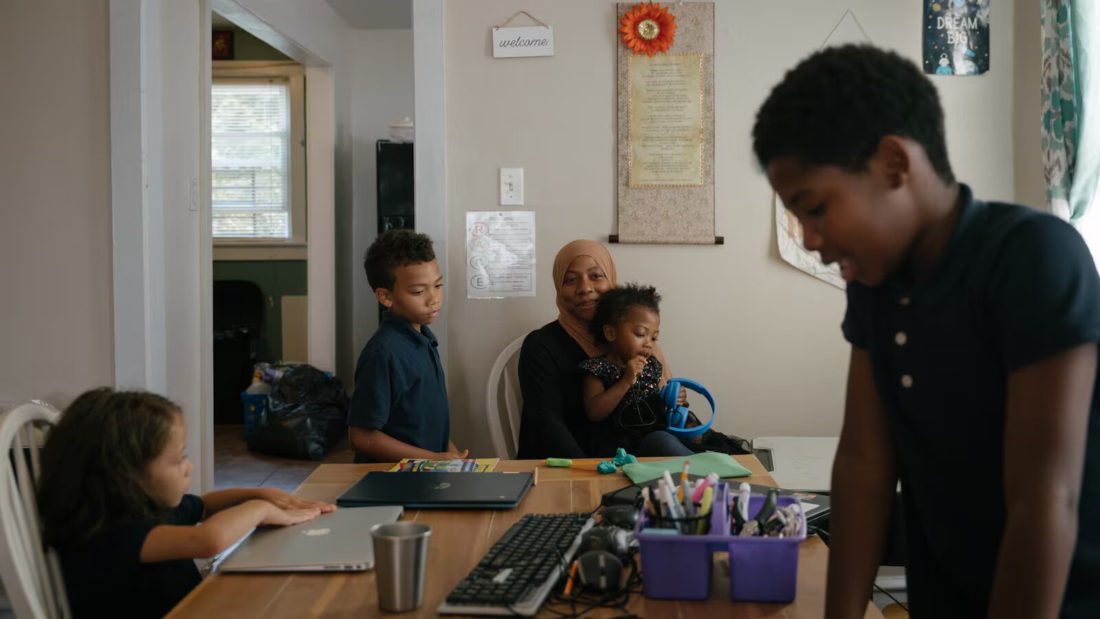 Online schools: 4 children with dark hair sit and stand around a dining table with school supplies in front of each of them while a woman in hijab leaning down next to one of them