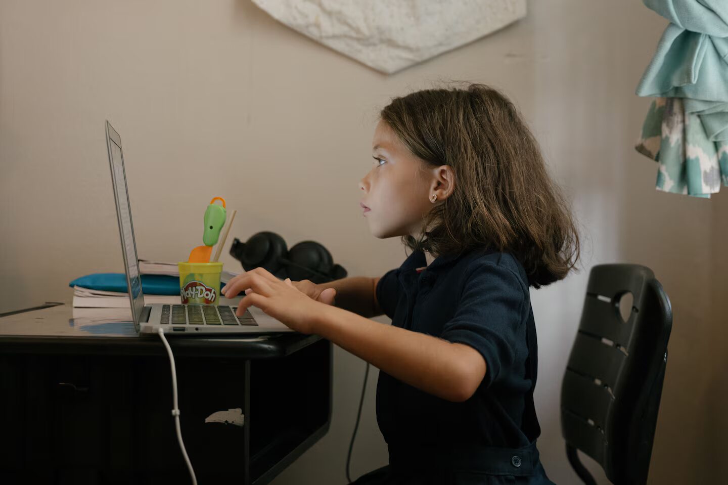 Online school: Side view of young girl with long, bobbed, dark hair sits at laptop lookinng at screen and typing on keyboard