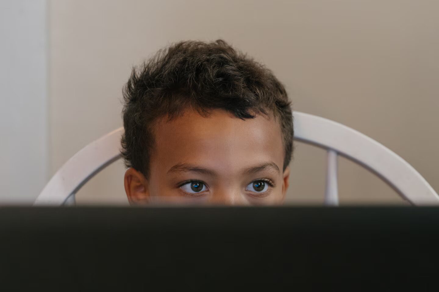 OOnline school: Top of head and eyes of young boy with short, dark curly hair looks at laptop screen in front of him