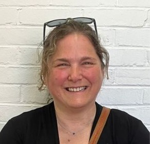 Competency-based innovative high school: Headshot of middle-age woman with light brown hair pulled back and black top in front of white brick wall.