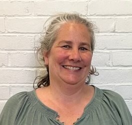 Competency-based innovative high school: Headshot middle-age woman with gray hair pulled back and sage green top in front of white brick wall.