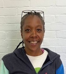 Competency-based innovative high school: Black woman with black hair pulled back in white top and dark vest in front of white brick wall.