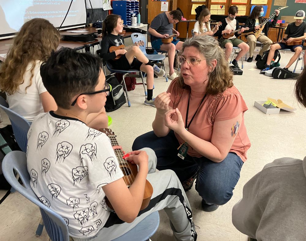 Competency-based innovative high school: Older, femaile teacher with gray hair in coral top and blue jeans crouches on floor in front of young, dark-haired teen boy in white shirt and gray sweat pants seated in chair holding ukelele