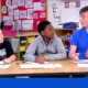 AMERICORPS TUTORS: YOUNG MAN IN BRIGHT BLUE AMERICOPRS POLO SHIRT SITS WITH TWO YOUNG BOYS IN CLASSROOM AT LONG DESK WRITING IN PAPER WORKBOOKS.