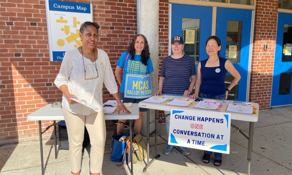 Mass. graduation requirements: Group of four ethnically diverse middle age women stand outside in a covered walkway at a table with info signs