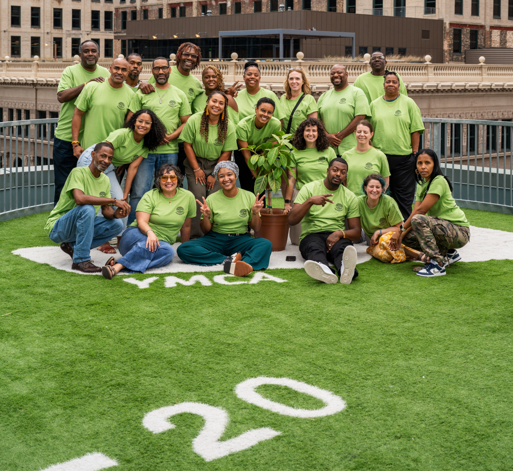 Restorative justice partnerships: Group of around 20 smiling people all wearing lime green t-shirts stand and sit in 3-4 rows on an artificial green turf field surrounded by tall city buidings