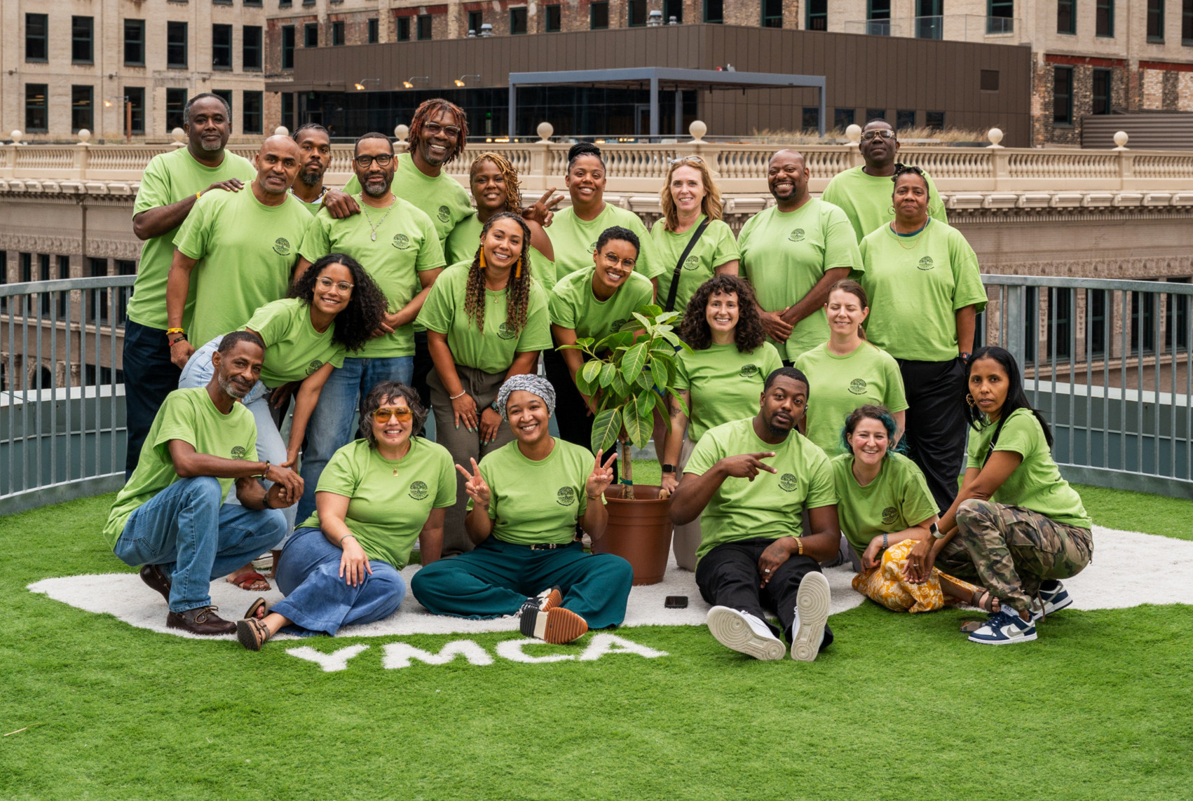 Restorative justice partnerships: Group of around 20 smiling people all wearing lime green t-shirts stand and sit in 3-4 rows on an artificial green turf field surrounded by tall city buidings