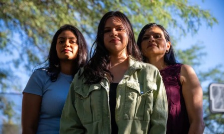 Latino gun reform Arizona: Thre Latino women with long, dark hair stand together facing camera with green tree foliage in the background