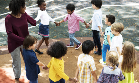 Childcare subsidies: Group of 8 primary students and one female teacher playing outside hold hands while going around in a circle