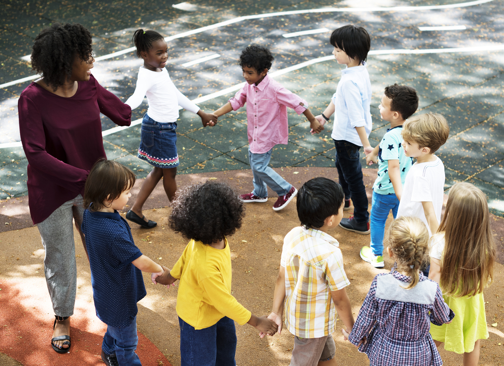 Childcare subsidies: Group of 8 primary students and one female teacher playing outside hold hands while going around in a circle