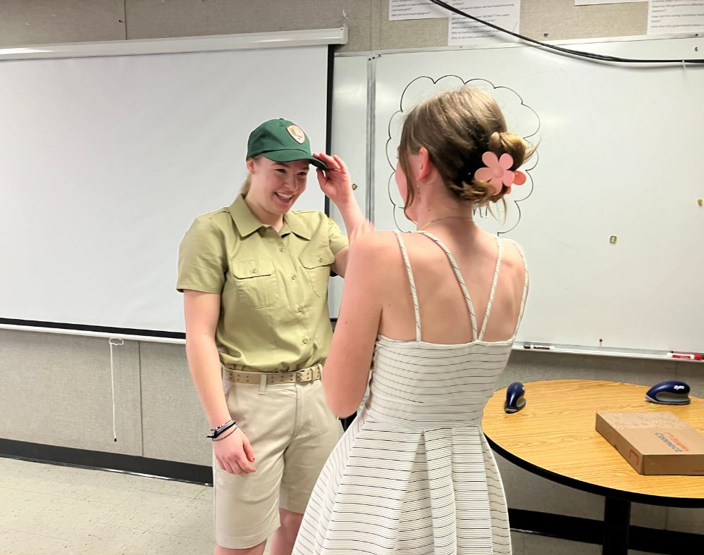 Competency-based innovative high school: Two teen girls face each other one in a park rangers tan uniform and one in a white dress