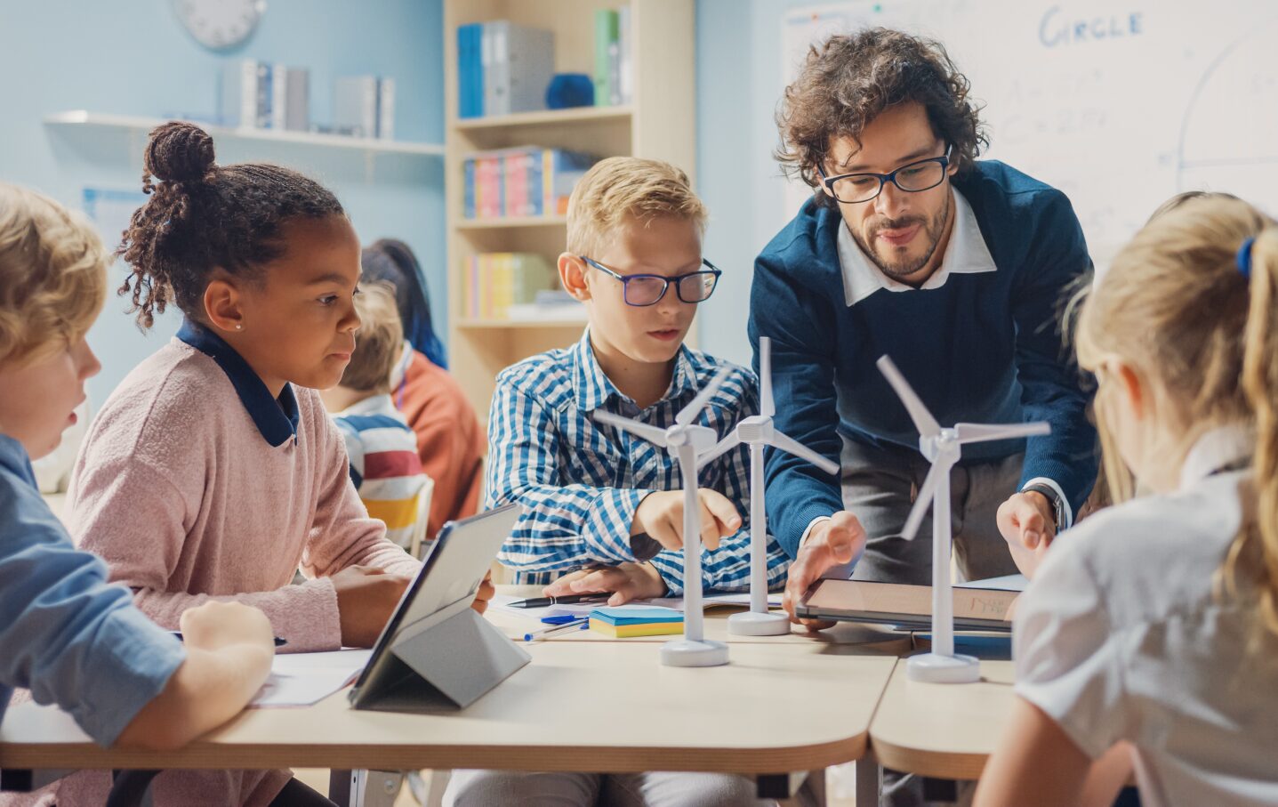 7 successful schools: Male teacher leans over table of 5 middle school students working on building models ofwind turbine power generators