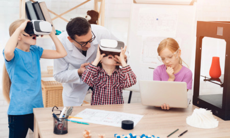 EdTech R&D: Elementary students in vr headsets and at laptop next to 3d printer sit and stand next to at desks while teacher lens in to help them