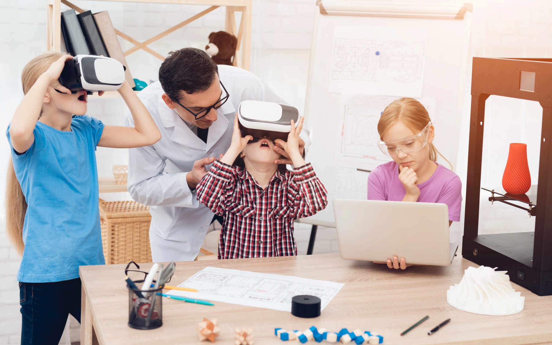 EdTech R&D: Elementary students in vr headsets and at laptop next to 3d printer sit and stand next to at desks while teacher lens in to help them