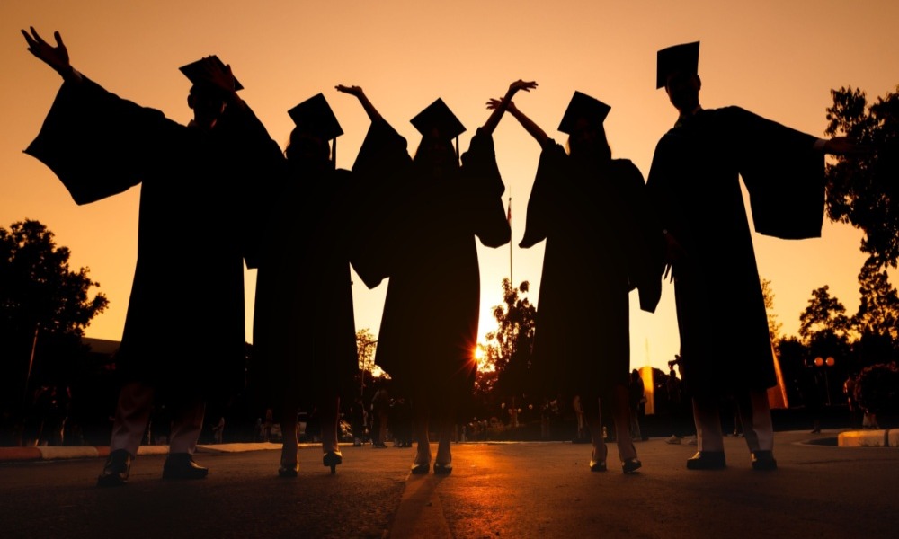 Dual enrollment: Black silhouette of 5 young adult in graduation gowns and caps standing with arms upraised against completely orange sunset sky