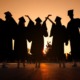 Dual enrollment: Black silhouette of 5 young adult in graduation gowns and caps standing with arms upraised against completely orange sunset sky