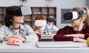 EdTech report: Happy elementary classmates looking at each other while sitting at desks gaming in vr headsets in classroom